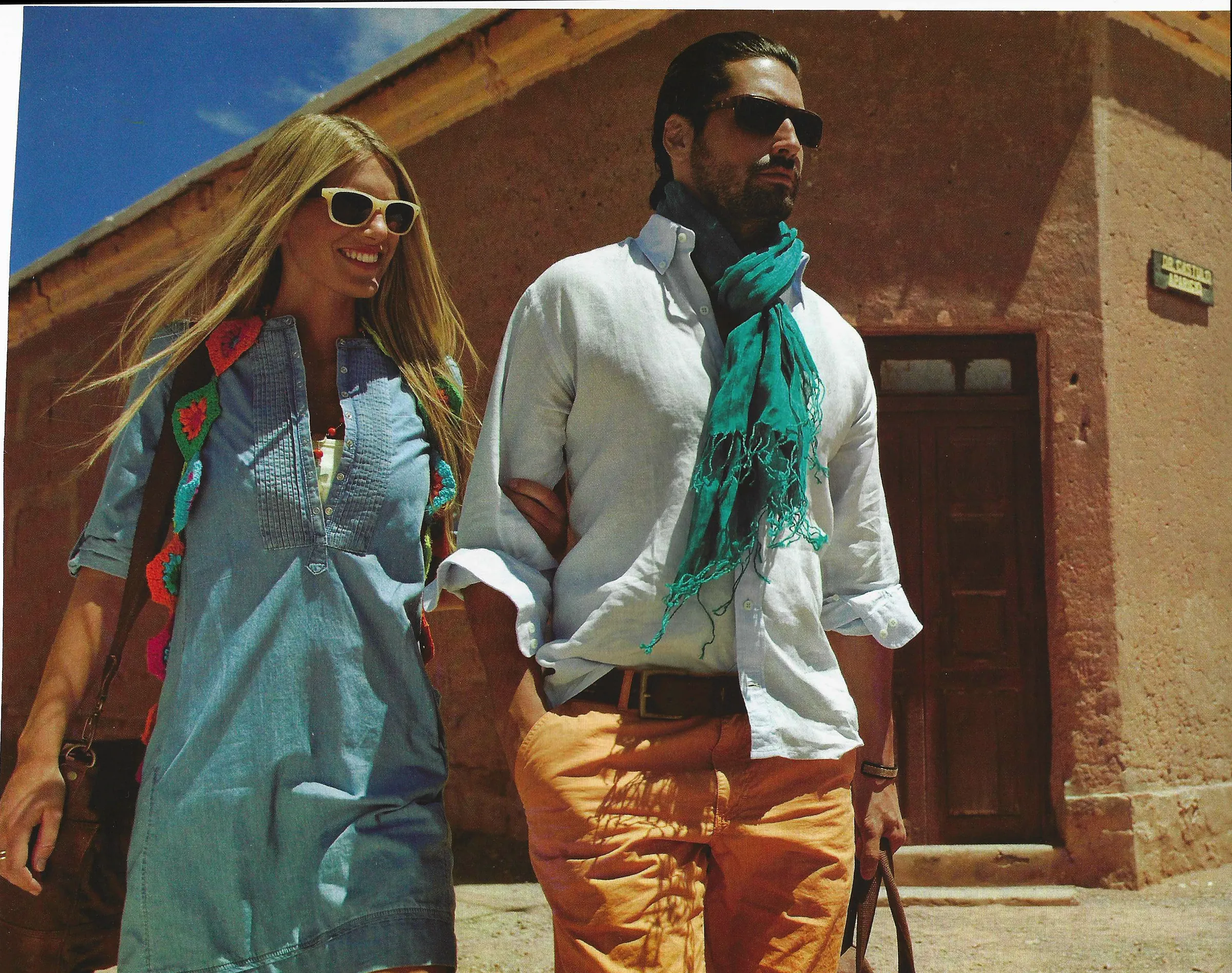 Nacho Valenti Lacroix in a white linen shirt and turquoise scarf, walking past an adobe wall in the sun