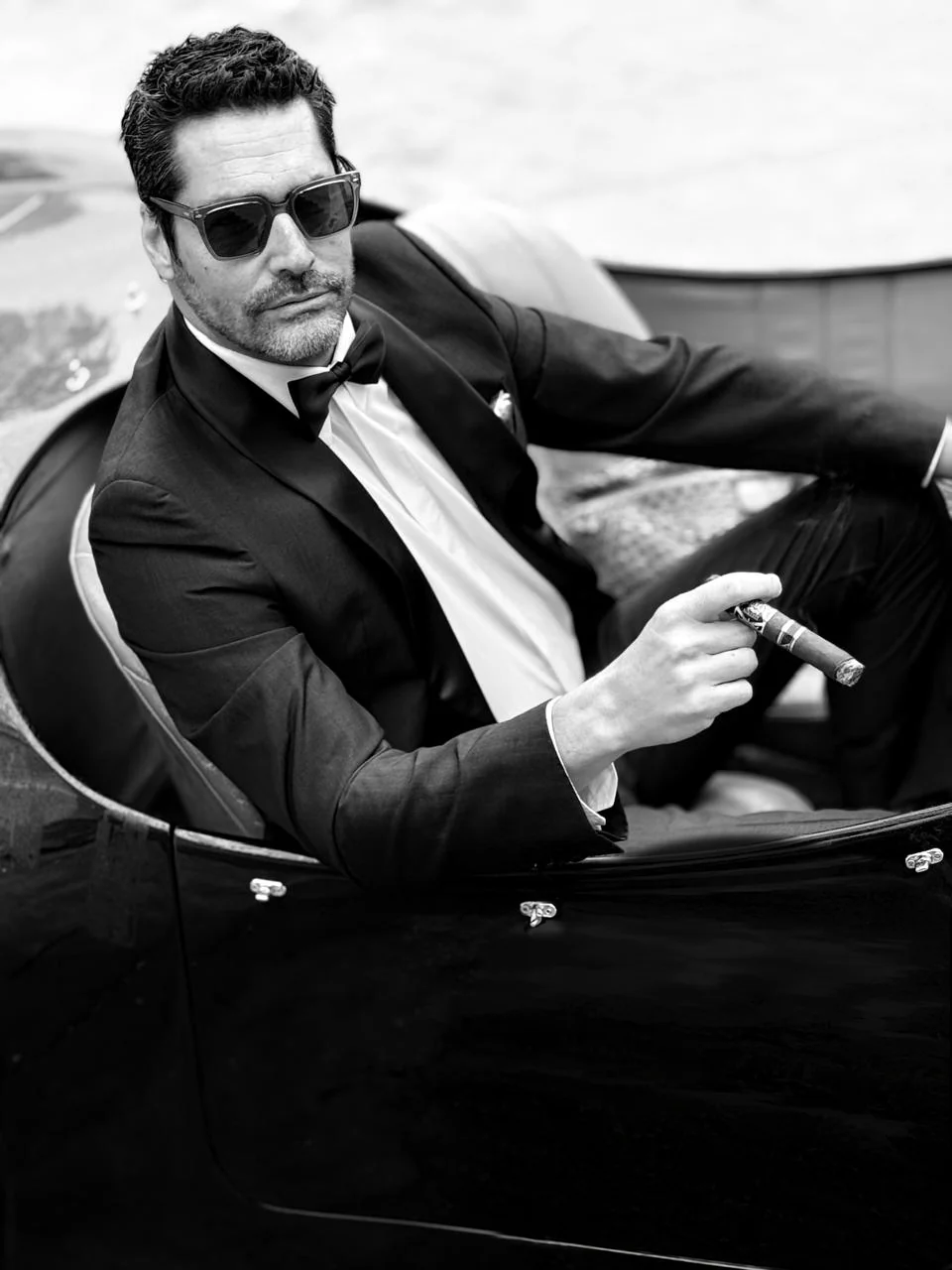 Nacho Valenti Lacroix in black tie and sunglasses, holding a cigar in an open vintage car, black and white