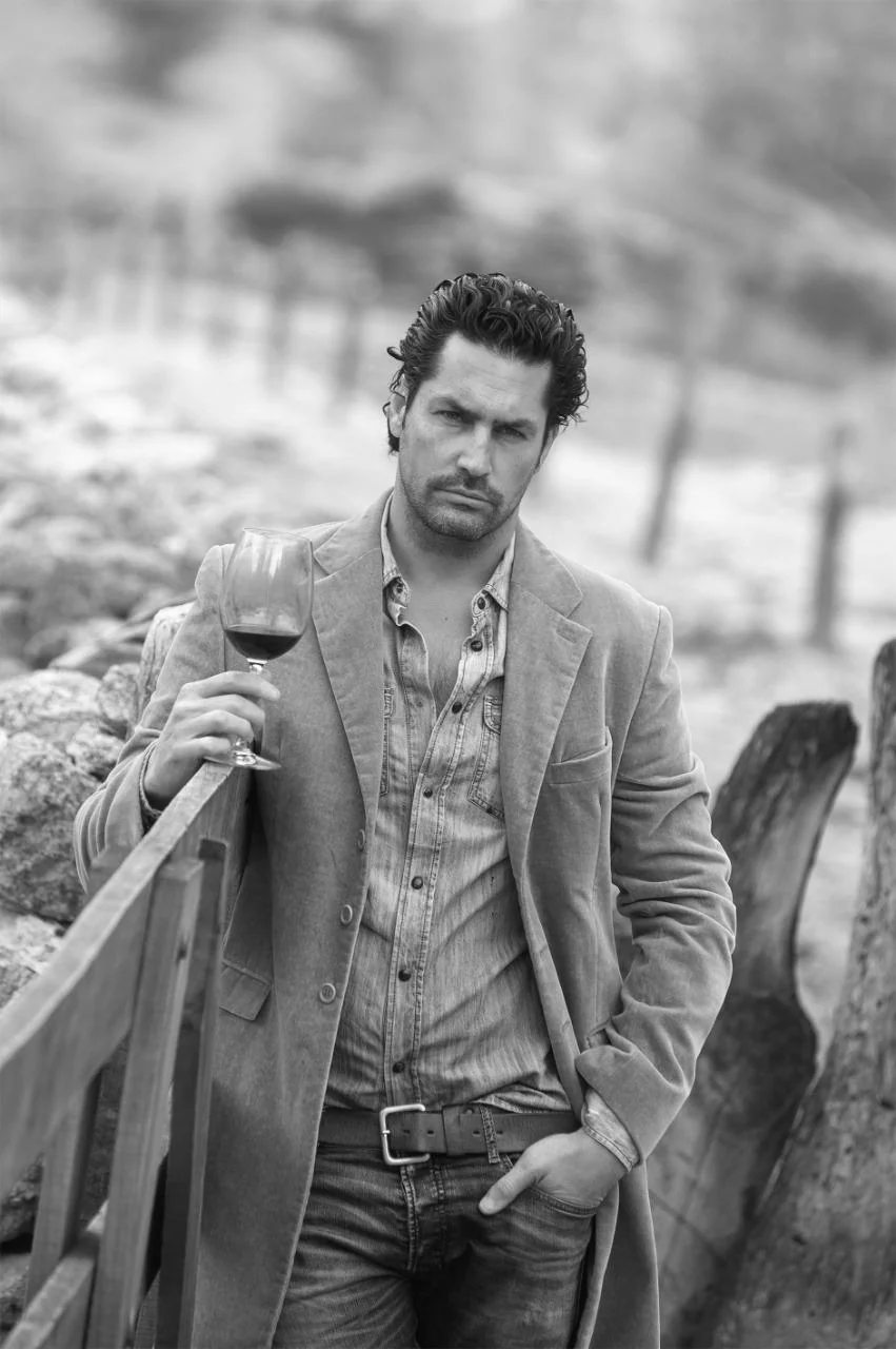 Nacho Valenti Lacroix leaning on a wooden fence at a winery, holding a glass of red wine, black and white